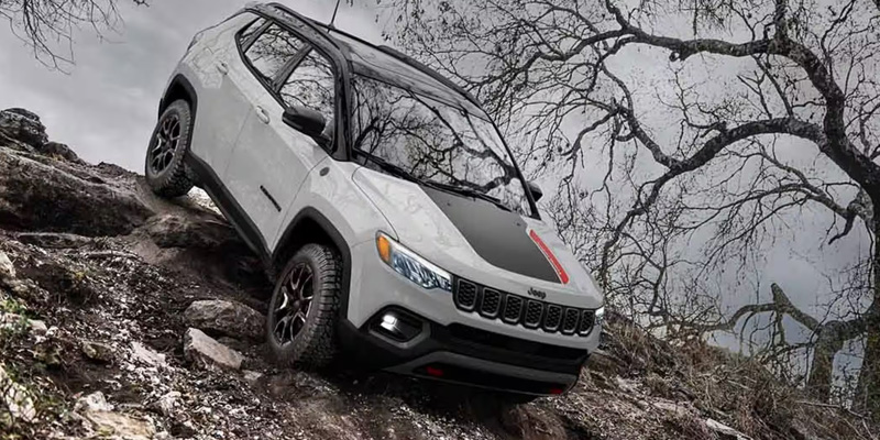 White Jeep Compass navigating rocky terrain in a rugged, off-road environment with bare trees in the background