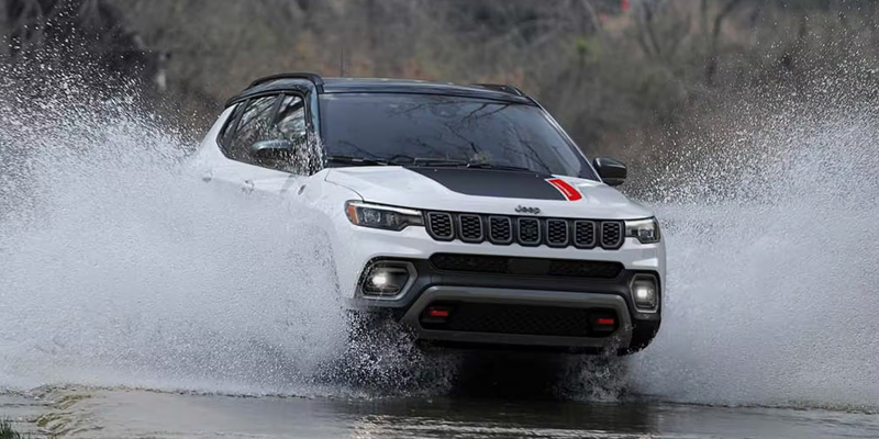 A white Jeep Compass Trailhawk with a black and red hood decal driving through a large puddle, creating a dramatic water splash on both sides of the vehicle.