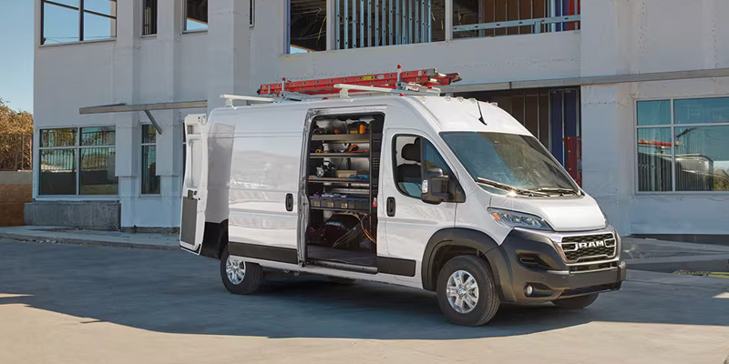 Front-three-quarter view of a white Ram ProMaster cargo van at a construction site with its doors open, showing storage shelves inside and roof-mounted ladders.