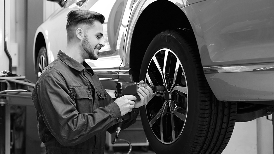 Black-and-white image of a mechanic using an impact wrench on a car wheel as the vehicle rests elevated on a lift inside a repair shop.