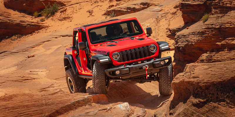 Front-three-quarter view of a bright red Jeep Wrangler Rubicon navigating a steep, rocky canyon trail in a desert landscape.